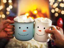 Close-up of two hands with snowman face on cup of hot cocoa with marshmallows. Mom and child relaxing together on a cozy winter evening by fireplace. Enjoy Christmas holidays, happy moments at home.
