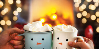 Close-up of two hands with snowman face on cup of hot cocoa with marshmallows. Mom and child relaxing together on a cozy winter evening by fireplace. Enjoy Christmas holidays, happy moments at home.
