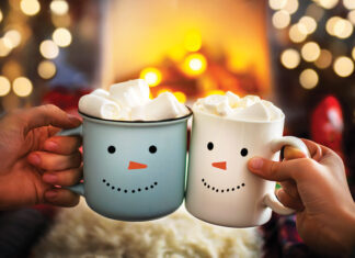 Close-up of two hands with snowman face on cup of hot cocoa with marshmallows. Mom and child relaxing together on a cozy winter evening by fireplace. Enjoy Christmas holidays, happy moments at home.