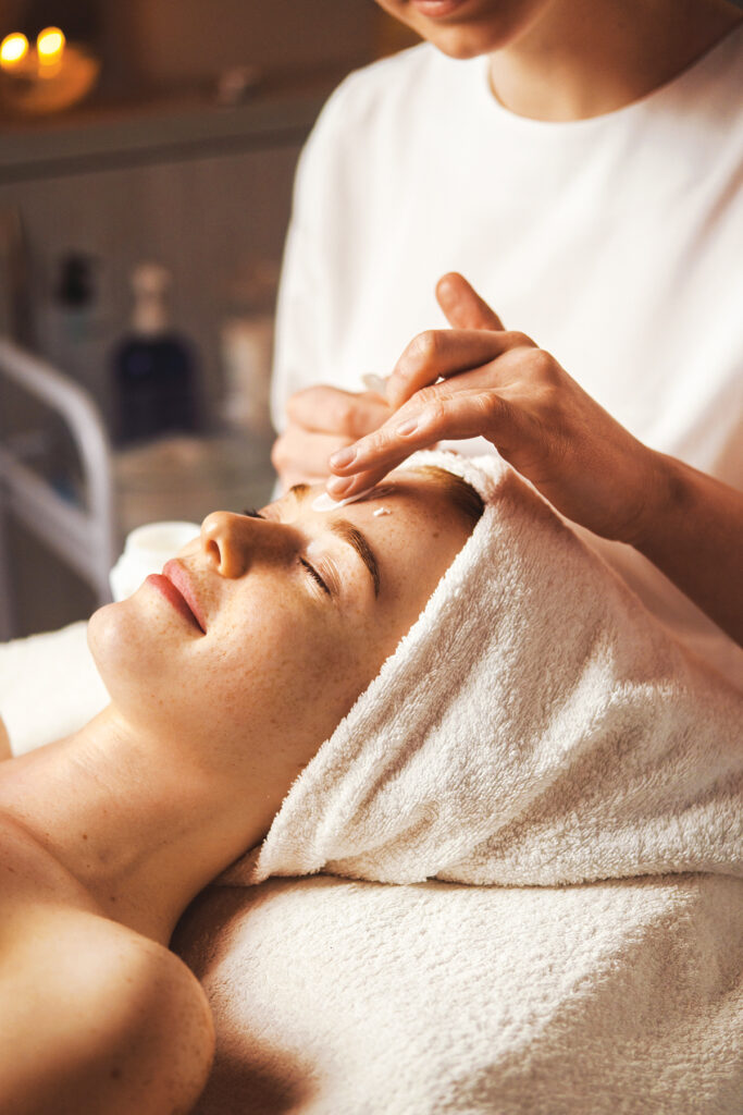 Beautician applying facial cream all over young woman's forehead at spa salon. Skin treatment. Girl facial treatment. Medical health.