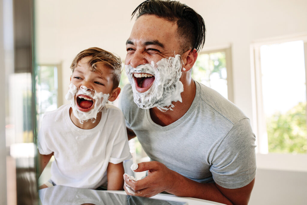 Man and little boy with shaving foam on their faces looking into the bathroom mirror and laughing. Father and son having fun while shaving in bathroom.