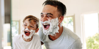 Man and little boy with shaving foam on their faces looking into the bathroom mirror and laughing. Father and son having fun while shaving in bathroom.