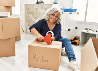 Middle age grey-haired woman smiling happy packing kitchen cardboard box at new home.