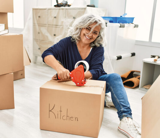 Middle age grey-haired woman smiling happy packing kitchen cardboard box at new home.