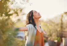 Young latin woman with arms outstretched breathing in fresh air during sunrise at the balcony. Healthy girl enjoying nature while meditating during morning with open arms and closed eyes. Mindful woman enjoying morning ritual while relaxing in outdoor park.