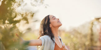 Young latin woman with arms outstretched breathing in fresh air during sunrise at the balcony. Healthy girl enjoying nature while meditating during morning with open arms and closed eyes. Mindful woman enjoying morning ritual while relaxing in outdoor park.