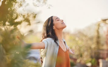 Young latin woman with arms outstretched breathing in fresh air during sunrise at the balcony. Healthy girl enjoying nature while meditating during morning with open arms and closed eyes. Mindful woman enjoying morning ritual while relaxing in outdoor park.