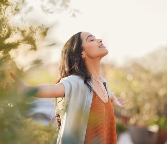 Young latin woman with arms outstretched breathing in fresh air during sunrise at the balcony. Healthy girl enjoying nature while meditating during morning with open arms and closed eyes. Mindful woman enjoying morning ritual while relaxing in outdoor park.